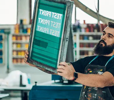 Man holding a screen printing frame in a colorful workshop, demonstrating screen printing techniques.